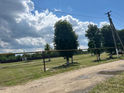 The image shows a rural scene in Dondukovskaya, Russia, under a partly cloudy sky with bright sunlight. The foreground is dominated by a dusty, unpaved road with some puddles reflecting the sky, suggesting recent rain. A low, rusty metal fence with a yellow top rail runs across the middle ground, separating the road from a grassy field.
Beyond the fence, several trees are visible, some with whitewashed trunks, a common practice in Russia to protect them from pests and the sun. In the background, there is a dark fence, some smaller buildings, and more trees, indicating a village or rural settlement. A utility pole with power lines stretches across the upper part of the image, suggesting the presence of electricity infrastructure.
The weather appears to be warm and fair, with dramatic cumulus clouds against a bright blue sky. The overall impression is one of a peaceful, everyday rural landscape, possibly in the late morning or early afternoon given the strong sunlight and shadows. There is no visible text in the image. The scene is devoid of people, focusing solely on the natural and man-made elements of the rural environment.
