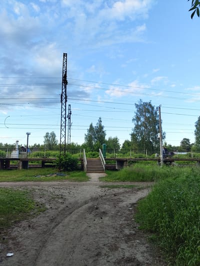 The image depicts an outdoor scene at a railway crossing in Tsibino, Russia. A raised pedestrian overpass with metal railings spans the railway tracks. To the right of the overpass, a set of concrete stairs with metal handrails leads up to it. Two individuals, one appearing to be an adult and the other possibly a child, are standing on the overpass, facing towards the right side of the image. In the foreground, a dirt path with tire tracks curves through overgrown grass. On the left side of the overpass, a white industrial-looking structure is visible behind the railing. Two tall, dark metal electrical pylons with numerous wires are prominent in the scene, one to the left of the overpass and another more centrally located. The sky above is blue with scattered clouds, and a dense line of trees forms the background. The overall impression is of a functional, albeit somewhat rustic, public crossing.