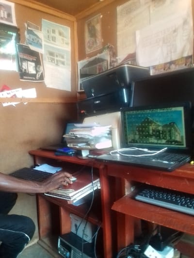 The image shows the interior of a small office in Auchi, Nigeria, located at coordinates 7.062 latitude, 6.273 longitude. The office appears to be a design or architectural firm. The walls are covered with drawings and blueprints. A man sits at a desk, using a computer. The desk is cluttered with papers, a printer, and a laptop. On the desk is a stack of papers and a computer with a drawing of a building on its screen. There are also multiple power cords and wires. The person is wearing a dark shirt and black pants. The office is small and cluttered, but it is functional.
