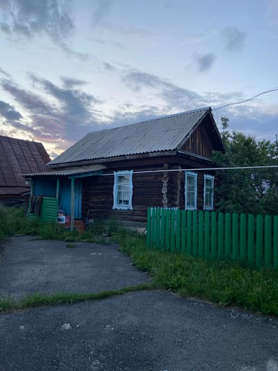 A rustic, weathered wooden house with a corrugated metal roof stands under a cloudy, twilight sky. The house features two visible windows with decorative white trim. A covered porch area with a bright blue door and green corrugated siding is attached to the front. A bright green wooden fence runs along the front of the property, separating it from a paved driveway with overgrown grass. Trees and foliage are visible behind the house and fence. The scene appears to be in a rural or semi-rural setting in Askarovo, Russia, likely a residential area given the presence of the house and fence. No people are present in the image.