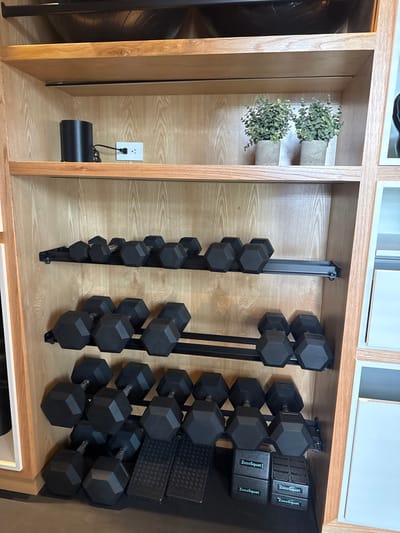 This image presents a detailed view of a multi-tiered wooden shelving unit used for storing fitness equipment within a facility in San Luis Potosí City, Mexico.
The shelving unit is constructed from light-colored wood, likely a veneer, and consists of at least four visible horizontal shelves, with the top section partially obscured.
On the second shelf from the top, a black cylindrical object is plugged into a white, dual electrical outlet centered on the back panel. To the right, two small artificial plants, each with green, leafy foliage, are housed in individual grey, concrete-textured pots. A black metal support bar runs along the underside of this shelf.
The third shelf down features a black metal two-tiered rack holding approximately eleven black, hexagonal-headed dumbbells. These dumbbells appear to be of lighter to moderate weights.
The fourth shelf down contains a similar black metal two-tiered rack, also holding approximately eleven black, hexagonal-headed dumbbells. These dumbbells are visibly larger than those on the shelf above, indicating heavier weights.
At the base of the unit, on the floor level, a third collection of black, hexagonal-headed dumbbells is present. Several are arranged on another black metal rack, while larger dumbbells rest directly on the floor or on two black, rectangular anti-slip mats with a diamond plate pattern. To the immediate right, four black rectangular blocks, each bearing the white text "ZocoSquat," are stacked vertically in two pairs.
The scene is static, depicting an organized storage area, with no individuals present or actions in progress. The equipment is uniformly black, contrasted by the natural wood tones of the shelving.