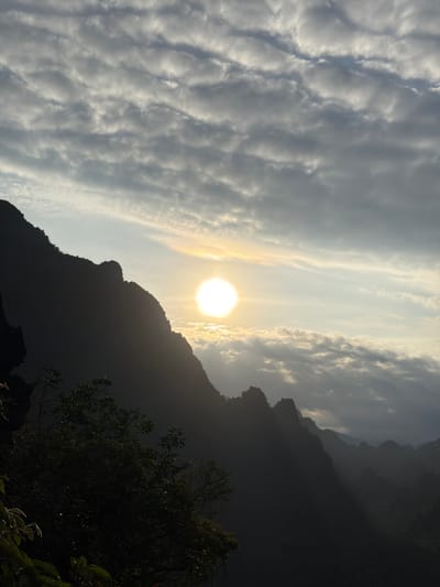 This image captures a dramatic sunset over a mountainous landscape, likely in the vicinity of Naka, Laos. The sun, a bright, luminous orb, is positioned slightly left of the center of the frame, casting a warm glow through a textured sky filled with layered clouds. These clouds exhibit a ripple-like pattern and are illuminated by the setting sun, creating a sense of depth and movement.

Dominating the left side of the image is a steep, dark mountain silhouette, its jagged peaks sharply defined against the lighter sky. Vegetation, appearing as dark masses of foliage, clings to the lower slopes of this mountain, with some leaves catching the light and appearing as small, bright glints. To the right of the sun, more distant mountain ranges fade into a hazy, bluish distance, suggesting a vast, expansive terrain.

The foreground is dominated by the dark, imposing mountain, which casts the lower part of the scene into shadow. The overall atmosphere is one of natural grandeur and tranquility, with the setting sun providing a focal point and a source of dramatic lighting. There are no people or animals visible in the image. The scene is entirely natural, showcasing the rugged beauty of the mountainous environment at dusk.