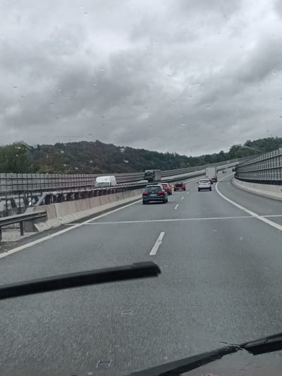 The image captures a view from inside a vehicle traveling on a multi-lane highway, likely the D1 highway, under overcast and rainy conditions. Water droplets are visible on the windshield, obscuring some of the view. The highway has clear lane markings and concrete barriers separating lanes and the roadside. Several vehicles, including cars and a white truck, are visible on the highway. The surrounding environment consists of green, tree-covered hills. The presence of a white truck with the number "11" on its rear suggests this location may be in the vicinity of Hvězdonice, Czechia, as this is a known service area on the D1 highway. The overall scene depicts a typical highway transit on a day with poor weather.