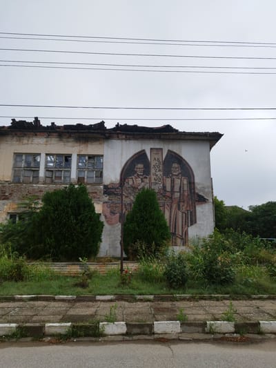The image shows a dilapidated building on a cloudy day. The building is partially ruined, with a damaged roof and broken windows. A mural of two figures in historical clothing is painted on the side of the building. In front of the building, there are two conical evergreen trees and overgrown vegetation. A sidewalk with a concrete curb separates the building from a paved road. The scene is located in Slanotran, Bulgaria.