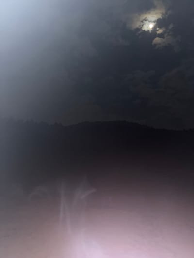 The image depicts an outdoor, low-light environment at night or under heavily overcast conditions in La Marquesa, Mexico. The upper portion of the frame shows a dark sky with significant cloud cover. A bright, diffused light source, consistent with a moon, is partially visible through gaps in the clouds in the upper right quadrant. Below the sky, a dark, silhouetted horizon line of what appears to be a forested mountain range or dense tree line extends across the midground. The lower half of the image is dominated by a diffuse, light-emitting haze, likely fog or ground-level mist. Within this haze, several elongated, upward-tapering luminous streaks are visible, resembling plumes of light reflecting through atmospheric conditions. There are no discernible people, specific objects beyond the natural landscape and atmospheric phenomena, or explicit actions or interactions depicted. The color palette transitions from dark blues and greys in the sky to pinkish-purple and white in the hazy foreground.