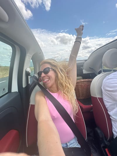 A blonde woman with curly hair, wearing sunglasses, a pink sleeveless top, and denim shorts, sticks her tongue out while riding in a convertible. Her right arm is raised in the air, adorned with several bracelets. She is seated in the front passenger seat of a car, with a seatbelt across her chest. The interior of the car features red and white upholstery. A wicker basket is visible in the back seat. The background shows a blue sky with white clouds and a rural landscape with a road. The image appears to be taken in Alaincourt, France, on a sunny day.