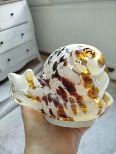 A hand is holding a glass decorative snail figurine. The snail is made of clear glass with swirling patterns of white, brown, and amber. It has a coiled shell and a pointed head with two antenna-like protrusions. The snail is resting on a white base, and the hand holding it is visible from the wrist down. The background shows a white dresser with black knobs, a white radiator, and a light-colored rug. The lighting suggests it is daytime, with natural light coming from the right. The overall scene appears to be an indoor setting, likely a room in a home. There is no visible text in the image.
