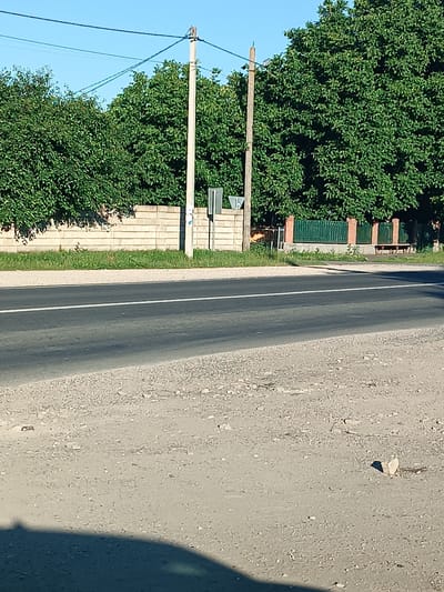 The image shows a view of a road in Copăceni, Moldova. The road has a white line marking the center. To the left of the road, there is a low, light-colored brick wall, topped with lush green trees. Two utility poles stand near the wall. Further to the right, a dark green metal fence with brick posts borders another area with trees and a wooden bench. The ground in the foreground is a rough, gravelly surface. The sky is clear and blue.