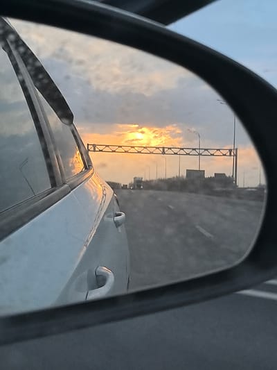 The image depicts a sunset view through the side mirror of a car traveling on a highway. The sun is positioned behind a metal structure spanning the road, with trucks visible further down the highway. The car's side is visible in the foreground, reflecting the sunlight. The location is inferred to be in Khutor, Russia, based on the metadata. The environment consists of a highway at sunset with overcast skies. There are no people visible in the image.
