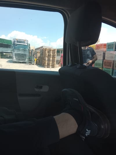 The image, taken from inside a vehicle, shows a view of a truck stop or loading area in Ανδραβίδα, Greece. A large Volvo truck is visible through the driver's side window, parked near stacks of wooden pallets and plastic crates. A forklift is positioned between the truck and the pallets. Two people are seen outside the vehicle, one wearing a red shirt and the other a dark t-shirt. The vehicle's interior is visible, including a headrest, and a leg with a black sock and a black Nike Air Max 270 sneaker rests on the dashboard. The sky is blue with some clouds.
