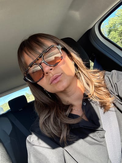 The image shows a close-up, selfie-style shot of a woman from the chest up, taken inside a car. She is wearing large, tortoise-shell framed sunglasses and a black collared shirt. A light-colored seatbelt is visible across her chest. Her hair is brown and extends past her shoulders. The background shows the interior of the car, including a headrest, and a blurred view through the window of green trees and blue sky, suggesting an outdoor, possibly rural, setting. There is no indication of the city of Mougins, France, in the image itself.