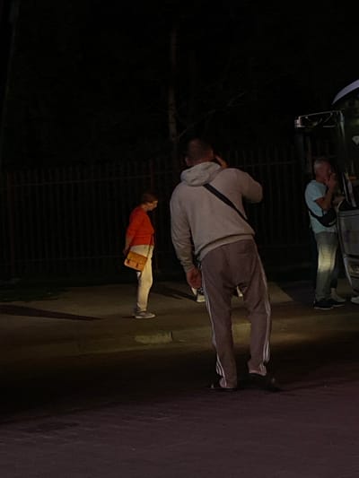 The image depicts a nighttime scene in Terespol, Poland, showing three individuals and a vehicle. The foremost subject is a person wearing a light gray hooded sweatshirt and dark pants with white stripes, standing and appearing to interact with something in their hand. Behind and to the left of this person, another individual is partially visible, wearing an orange top and white pants, carrying a light brown bag. Further in the background, a third person, dressed in a light blue shirt and gray pants, is seen near the back of a vehicle. The vehicle itself is dark and appears to be parked. The lighting suggests it is nighttime, with artificial lights illuminating the scene. The ground appears to be paved. No visible text is present in the image.