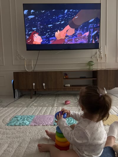 This image depicts an indoor domestic scene in Velikovechnoye, Russia, likely a living room. A young child, viewed from behind, is seated on a light-colored shag rug, actively playing with a multi-colored stacking ring toy. The child has light brown hair tied in a ponytail with a purple elastic and wears a white long-sleeved top with a faint floral pattern. Partially visible behind the child's right leg is another person's leg, clad in blue jeans and a white sock.
In front of the child, on the rug, are several interlocking textured mats in shades of light blue and purple. To the child's right, a deflated pink heart-shaped balloon rests on the rug.
Dominating the upper half of the image is a large, wall-mounted flat-screen television displaying a scene from an animated movie. The scene features a female animated character with red hair looking upward, while a male animated hand reaches down toward her against a dark, snowy background with visible architectural elements.
Below the television is a dark wood entertainment cabinet with vertical ribbed paneling and open shelves. Two white child safety locks are visible on the cabinet's central doors. A white PlayStation 5 console stands vertically on the far left of the cabinet. Various decorative items are placed on the cabinet: a white U-shaped candle holder with two white candles on the left, a green mushroom-shaped glass object, and two tall white taper candles in gold holders on the right. The wall behind the television and cabinet is light-colored and features white decorative molding, including ornate scrollwork. The floor beneath the rug appears to be a light-colored tiled surface.