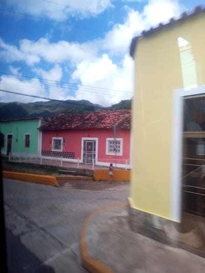 The image depicts a street scene in Tacarigua, Venezuela, featuring colorful buildings and a view of mountains in the background. In the foreground, a blurred asphalt road curves to the right, bordered by a yellow curb. A person, appearing to be a child, is visible in the middle ground, standing on the sidewalk in front of a red building. This building, along with an adjacent green building, has traditional tiled roofs and white-trimmed windows and doors. A large, pale yellow building dominates the right side of the frame, obscuring part of the view of the other structures. Overhead, power lines crisscross against a bright blue sky with scattered white clouds. The overall impression is of a sunny day in a tropical or sub-tropical town.