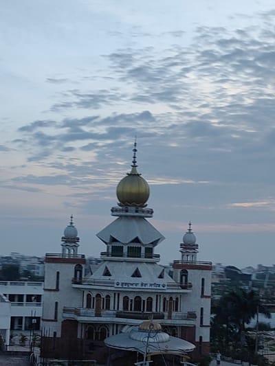 The image depicts a religious building, likely a Gurdwara, in the city of Baldhi, India. The structure features a prominent golden dome atop the central section, flanked by two white minarets with smaller domes. The building has multiple levels, white walls with red accents, and arched windows. In the background, the cityscape of Baldhi is visible, with hints of residential and commercial buildings. The sky is overcast with scattered clouds, suggesting it might be early morning or late evening. The overall scene conveys a sense of peace and architectural grandeur.