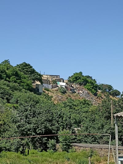This media file shows an outdoor scene featuring a hillside dotted with buildings and dense vegetation. The sky is a clear, bright blue, suggesting a sunny day.

The hillside is covered in green trees and bushes, with some areas of dry, brown earth. Several houses are visible, appearing to be a mix of weathered structures and more modern buildings. Some of these are clustered together, while others are more spread out. One notable area on the hillside appears to be a large accumulation of trash and debris.

In the foreground, there is more greenery, with a fence or railing made of rusty metal pipes. A stone wall and a wooden post are also visible. The overall impression is of a semi-rural or informal settlement built on a sloped terrain. There are no people or obvious activities taking place in the frame. The time of day appears to be daytime due to the bright sunlight.