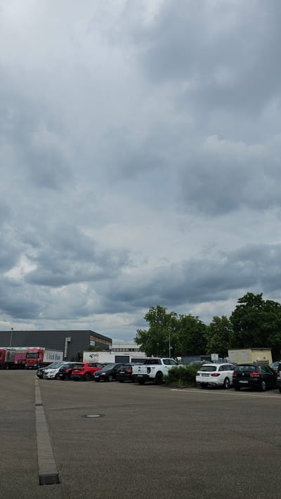 The image shows an outdoor parking lot filled with various vehicles, including cars, a white pickup truck, and several red trucks. The parking lot is paved with asphalt and has distinct demarcation lines. In the background, there are industrial buildings, a few trees, and a cloudy sky. One of the trucks has "Ulrich Haus" written on its side. The overall scene suggests a commercial or industrial area. Based on the presence of German license plates and the general architecture, it is likely located in a German city. The specific location is a parking lot in Neu-Ulm, Germany.