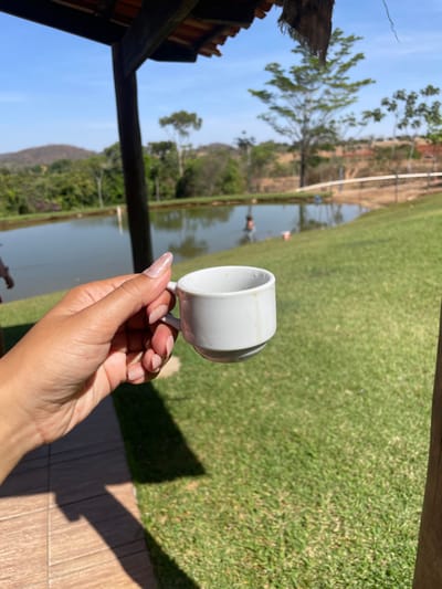 The image provides a slightly low-angle view of a right human hand holding a small, white ceramic cup with a handle, in Pirenópolis, Brazil. The hand, exhibiting manicured nails with a light, French-tip style, is in sharp focus in the foreground. The cup shows visible light brown stains on its exterior. Below the hand, a section of light brown square-tiled flooring is visible, partially shadowed, transitioning to a bright green grass slope. A dark wooden support post or pillar frames the right side of the foreground. Above, a wooden roof structure with reddish-brown tiles casts shadows.
In the midground, a dark green pond occupies the central and left portions of the frame. At least two individuals are partially submerged in the pond; one figure is visible on the left, and another is positioned towards the right with their back to the viewer. A white, cylindrical object, possibly a marker or buoy, floats in the water to the left. The background consists of rolling green hills densely covered with various trees and vegetation, under a clear, cloudless blue sky. The scene is illuminated by strong sunlight, indicative of an outdoor, natural environment.