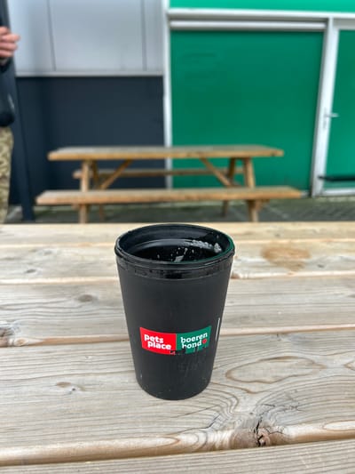 A black thermal mug is prominently featured in this image, resting horizontally on a light-colored wooden surface, likely an outdoor table or deck. The mug's opening is visible, showing a small amount of foamy liquid inside. On its side, two distinct logos are displayed: "Pets Place" in red text on a white rectangular background with a paw print graphic, and "boeren bond" in white text on a green rectangular background, with a red heart icon replacing the 'o' in 'bond'.
In the background, a large green wall or panel is visible, with a white frame along its top edge. Below this green section, there are darker grey and white panels, suggesting the exterior of a building. Further left and slightly blurred, a wooden structure, possibly part of a bench or outdoor furniture, can be seen. In the bottom-left corner of the frame, the fingers of a person's hand are partially visible, suggesting human presence nearby. The scene is brightly lit by natural light, indicating it is daytime. The combination of the wooden surface and background elements points to an outdoor setting in Ede, Netherlands.