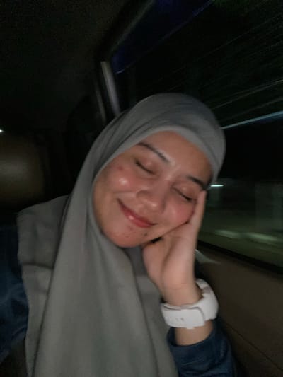 The media file is a close-up selfie of a young woman wearing a grey hijab and a dark blue shirt. She is lying down, with her eyes closed and a relaxed smile on her face, her left hand resting on her cheek. Her skin appears to have a dewy sheen, possibly from sweat or skincare products, and there are some visible blemishes. A white smartwatch is visible on her wrist. The background is dark and out of focus, with hints of green and blue light suggesting an outdoor or dimly lit indoor environment, possibly at night. The overall mood is peaceful and intimate. The context provided indicates the location is Jawa, Indonesia.