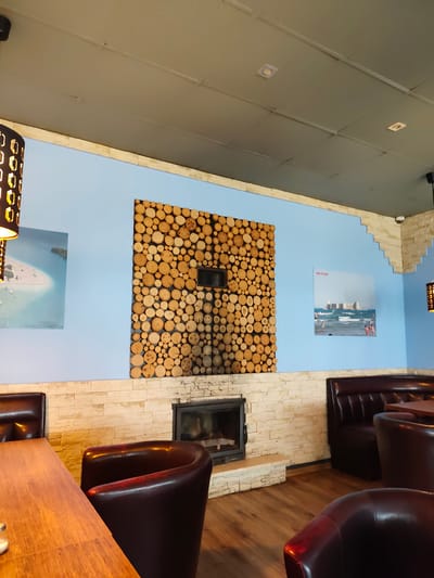 This image depicts the interior of a restaurant with a light blue wall as the backdrop. On the wall, there is a square arrangement of circular wood slices above a fireplace. To the left of the wood arrangement, a framed picture shows a beach scene with turquoise water and white sand. To the right, another framed picture displays a castle on a body of water with people in the foreground. The room is furnished with dark brown leather booth seating and individual chairs. A wooden table is partially visible in the foreground. The ceiling is a neutral gray with recessed lighting fixtures. The overall ambiance suggests a modern and cozy dining space. This restaurant is located in Zgierz, Poland.