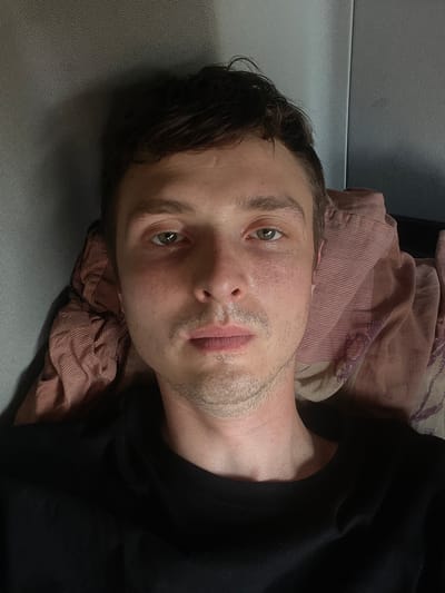 A close-up, low-angle, outdoor shot captures a young man lying on his back, looking up at the camera. He is wearing a black t-shirt. His head is tilted to his left, and his eyes are open, with a slightly weary expression. The man has light skin with visible freckles and stubble on his chin and jawline. His hair is dark brown and appears slightly tousled.

He is positioned on a gray surface that could be a bench or the ground, with a patterned, pinkish-brown fabric laid behind and beneath his head, possibly a blanket or clothing. The background is blurred, but suggests an outdoor, possibly urban or semi-urban environment, given the hints of textured surfaces and distant objects. The lighting is somewhat dim, suggesting it might be evening or a cloudy day. No text is visible in the image. The overall mood is candid and informal.