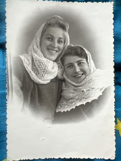 This is a black and white photograph of two young women, likely friends or sisters, posing together. They are both wearing headscarves with intricate lace patterns. The woman in the foreground is looking directly at the camera with a bright smile, her hair styled in soft waves. The woman behind her is also smiling and looking towards the camera, her face partially obscured by the headscarf. They appear to be embracing or leaning into each other, creating an intimate and affectionate composition. The background is softly blurred, focusing attention on the subjects. The photograph has a vintage feel, suggested by the style of dress and the black and white format. The edge of the photograph has a decorative scalloped border. The image is placed on a textured blue surface, possibly fabric, with a hint of a yellow and green pattern visible at the top right corner. The overall mood is warm and joyful. There is no visible text in the image. The context provided is "Дружба Снт, Russia", which suggests a location in Russia.
