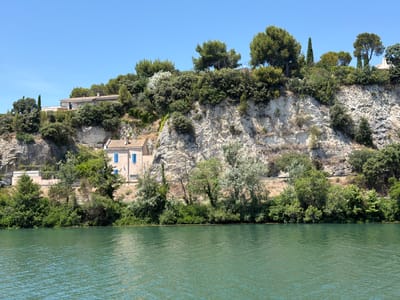 The image shows a wide, calm river with turquoise water in the foreground. On the far bank, a steep, rocky cliff rises, covered with lush green vegetation and scattered trees. Several houses are visible nestled into the hillside. One house, closer to the river and painted a light color with blue shutters, has a terracotta roof. Further up the slope, another building with a similar roof is partially visible, surrounded by trees. The sky above is a clear, bright blue. The scene is one of natural beauty and residential development along a riverbank, likely in a Mediterranean climate given the vegetation. This location appears to be on the Rhône River in Avignon, France, as evidenced by the characteristic landscape and architecture found in the area.