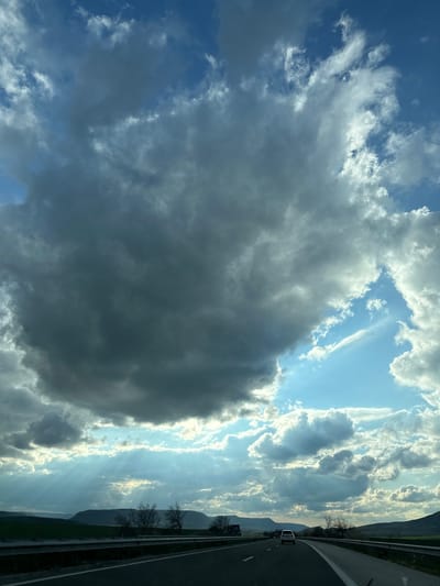 The image is a landscape oriented shot taken from a moving vehicle on a highway, presumed to be near Mlada Gvardia, Bulgaria. The left side of the image is dominated by a large, dark cumulonimbus cloud formation filling a substantial portion of the sky. The sky behind and around the cloud displays patches of blue interspersed with various other types of clouds. Sunlight appears to be streaming through some of the cloud formations, creating a visible light ray effect. The right side of the image depicts a highway with a guardrail separating the road from a green field and distant mountains. A single vehicle is visible on the highway, moving in the same direction as the camera. Bare trees are visible along the road.