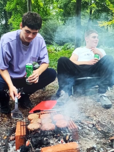 The image portrays two young males conducting an outdoor cooking activity in Aktash, Uzbekistan. The male on the left, possessing dark hair and clad in a light purple oversized t-shirt, dark trousers, a silver chain, and a ring on his right hand, is in a squatting position. He holds a green beverage can, identifiable as Heineken Original, in his left hand and a silver metal spatula in his right, manipulating food on a grill. The male on the right, with short light-colored hair and wearing a white t-shirt with a dark logo and dark trousers, is seated on a dark portable folding chair. He also holds a green beverage can (Heineken Original) in his right hand and is looking towards the right with his mouth slightly open.
In the foreground, a makeshift grill is assembled, consisting of a metal grate supported by two orange bricks, under which smoldering embers and charcoal generate smoke that rises vertically. Approximately seven hamburger patties, appearing raw or partially cooked, are arranged on the grate. A red flat object, possibly a cutting board, is situated behind the grill. The ground surface comprises dry grass, dirt, and scattered organic debris. The background reveals a dense arrangement of green trees and foliage, indicative of a wooded or forested environment under daylight conditions.