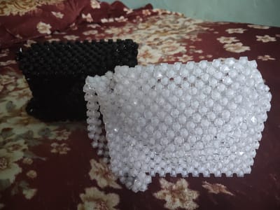 Two beaded bags, one black and one white, rest on a floral-patterned fabric, likely a bedspread. The black bag is to the left and appears to be made of smaller, faceted cubes. The white bag is larger, to the right, and is constructed from clear, faceted beads that catch the light. The fabric is a deep maroon with scattered cream-colored floral motifs. The background is slightly out of focus, suggesting a shallow depth of field. The lighting is soft, indicating an indoor setting.