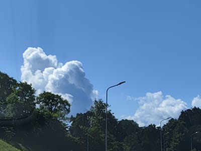 In the city of Mandegas, Latvia, a scene unfolds under a vast, clear blue sky dotted with voluminous white clouds, characteristic of a bright summer day. The foreground is dominated by a dense, dark green treeline, obscuring much of the ground. The trees are lush and full, with varying shades of green suggesting a variety of species.
Positioned prominently against the sky are two streetlights. The taller one, on the right, features a sleek, modern design with a curved arm extending over the roadway. Its dark metal pole stands tall, a stark contrast to the vibrant blue of the sky and the soft white of the clouds. To its left, a second, shorter streetlight is partially visible, also appearing to be a modern design.
The clouds are a significant element, with a large, cumulus cloud formation dominating the left side of the frame. Its puffy, textured surface suggests considerable height and volume. Smaller, wispy clouds are scattered across the sky, adding depth and dynamism to the overall composition. A faint, diagonal line suggesting a grassy slope can be seen on the far left of the image, partially obscured by the trees. There are no people or animals visible in the image.