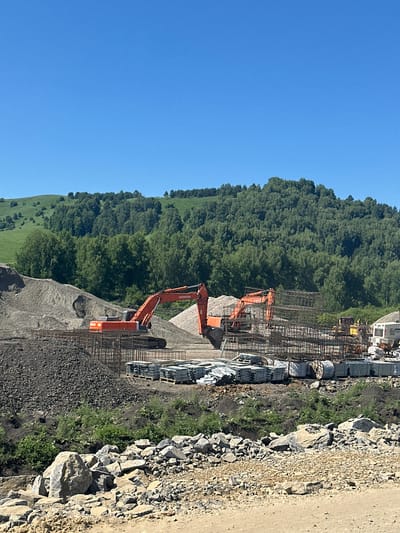In Altaiskoe, Russia, a construction site is depicted under a clear blue sky. The foreground is dominated by piles of loose gravel and large rocks, with smaller green vegetation growing among them. In the midground, two large orange excavators are positioned. The excavator on the left is in the process of digging into a pile of grey gravel. The excavator on the right is also actively engaged, with its boom extended. In front of the excavators are stacks of metal rebar cages and bundles of concrete slabs, all neatly arranged on pallets. Further in the background, a white construction vehicle is visible. The site is bordered by a lush green hillside covered with trees, with a distant rolling green hill under the vast expanse of the sky.