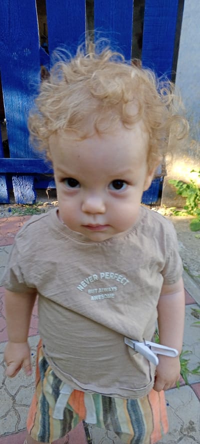 A toddler with blonde curly hair stands in front of a bright blue fence, looking directly at the camera with a serious expression. The child wears a light brown t-shirt with the text "NEVER PERFECT BUT ALWAYS AWESOME" printed on it in white, and striped shorts in shades of orange, green, and yellow. A white clothespin is attached to the side of the t-shirt. The ground beneath the child is paved with interlocking stones. Green foliage is visible to the right of the child and behind the fence. The lighting suggests it is daytime, possibly late afternoon due to the shadows. The scene appears to be outdoors, in a yard or garden setting in Novo-Tsimlyanskaya, Russia.
