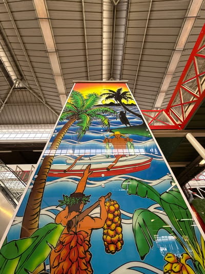 The image displays a tall, tapering column, approximately 8 meters in height, situated within a large indoor facility. The column is covered with a multi-paneled mural depicting Tahitian cultural scenes.

The mural features three main sections:
1.  **Top:** A sunset scene with an orange-yellow gradient sky, two silhouetted palm trees on a dark landmass, and dark blue water with white waves. A small, dark, indistinct figure is visible near one palm tree.
2.  **Middle:** A bare-chested male figure wearing a yellow head covering stands waist-deep in blue water, holding two fish. Behind him, a red outrigger canoe, labeled "TAHITI" on its side, is visible with paddles.
3.  **Bottom:** A male figure, viewed from behind, wears a leafy head lei and a skirt of banana leaves. He carries a large bunch of yellow bananas suspended from a pole over his right shoulder. Large green banana leaves are depicted on either side. The background is blue water with white waves.

The surrounding environment includes a corrugated metal roof structure with exposed beams. To the left of the column, an ascending escalator with metallic handrails and red side panels is visible. To the right, a prominent red, triangular lattice-work support structure extends upwards. The mural's surface is reflective, showing distorted reflections of the overhead structures, and is secured by circular fasteners.

This scene is located within a public building in Papeete, French Polynesia, a collectivity of France, likely Faa'a International Airport given the architecture and explicit Tahitian branding.