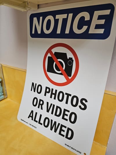 A white sign with black text and a red circular symbol of a camera with a slash through it is displayed. The text on the sign reads "NOTICE" in large blue letters at the top, followed by "NO PHOTOS OR VIDEO ALLOWED" in large black letters. The bottom of the sign includes a website address "SignMission.com", a phone number "1-561-508-6513", and a product code "OS-NS-P-1014-V-14753". The sign is attached to a light-colored wall and is leaning against a yellow surface. The environment appears to be indoors, possibly a retail or public space, given the nature of the sign and the surrounding elements. No people are visible in the image.