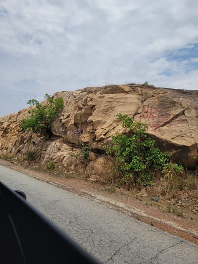 A photograph taken in Rimin Ziem, Nigeria, shows a large, tan-colored rock formation adjacent to a cracked asphalt road. The rock face is marked with reddish-pink graffiti and interspersed with green vegetation and dry grasses. A partially obscured section of what appears to be a dark car interior is visible in the lower right corner of the frame. The sky above is visible and is filled with scattered clouds.