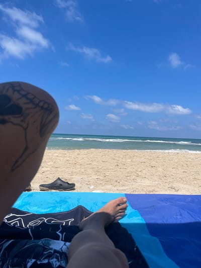 A first-person perspective shot captures a person relaxing on a beach in Ga'ash, Israel. The image focuses on the person's leg with a skull tattoo on their thigh, and their bare foot resting on a blue beach towel. A black sandal is placed on the sand nearby, with the ocean and sky visible in the background. The sea is calm with gentle waves, and the sky is bright blue with scattered white clouds, indicating a sunny day. The sand is light brown and appears dry.
