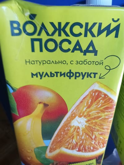 The image shows the front of a juice carton, likely a Tetra Pak style, with a bright yellow background. Dominating the top half of the carton are large, dark blue Cyrillic letters. The top line reads "ВОЛЖСКИЙ" and the second line reads "ПОСАД". Below this text, in smaller, lighter blue Cyrillic letters, is the phrase "Натурально, с заботой" which translates to "Naturally, with care". Further down, in even larger, dark blue Cyrillic letters, is the word "МУЛЬТИФРУКТ", meaning "Multifruit". To the right of the "МУЛЬТИФРУКТ" text is a decorative swirl that ends in an arrow pointing downwards.
The lower half of the carton features realistic illustrations of fruit. On the left, there is a close-up of a red fruit, possibly an apple or peach, with a stem. To its right and slightly in front is a yellow banana with visible water droplets on its peel. The right side of the carton shows a large slice of orange, revealing its segments and texture. The colors of the fruit are vibrant, with red, yellow, and orange tones.
The overall presentation suggests a commercial product, likely juice, emphasizing its natural ingredients and fruit content. The image does not contain any information that would allow for its location to be determined within the city of Velykyi Sukhodil, Ukraine, or any other specific location.