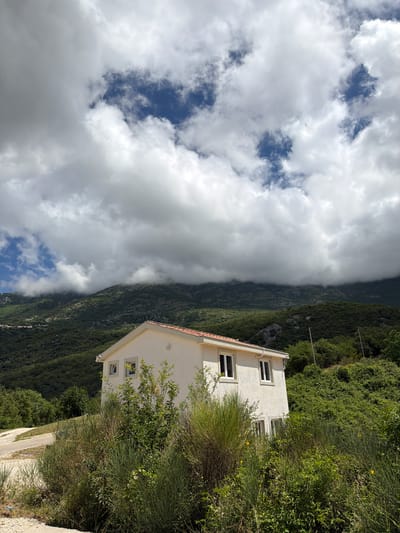The image depicts a serene mountainous landscape in Bratešići, Montenegro, under a partly cloudy sky. In the foreground, a modern two-story white house with a reddish-brown tiled roof is partially obscured by dense, wild green vegetation, including tall bushes and trees. Several windows are visible on the house's facade. A light-colored dirt or gravel path runs along the bottom edge of the frame, leading towards the house.
Behind the house and extending across the midground, verdant mountains rise steeply, covered in a thick canopy of trees and foliage. A dramatic band of low-hanging, dark grey clouds stretches horizontally across the mountain range, partially obscuring the peaks and creating a sense of depth and atmosphere. Above these lower clouds, the sky features a mix of bright white and softer grey clouds, with distinct patches of clear blue sky visible.
The scene captures a peaceful, natural environment, with no discernible human activity or events taking place. The lighting suggests it is daytime, possibly morning or early afternoon, with ample natural light illuminating the landscape. The weather is dynamic, showcasing both clear skies and significant cloud cover. There is no visible text in the image.