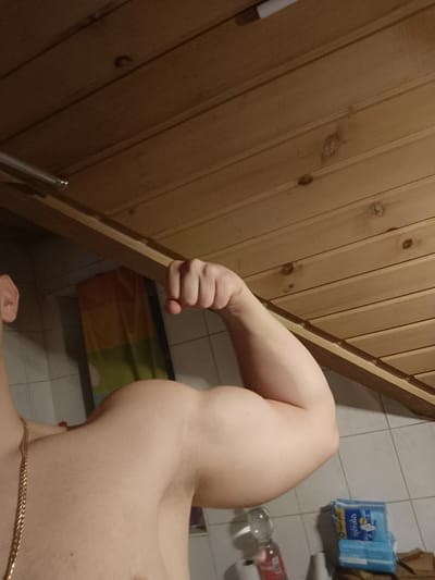 The image shows a muscular arm flexing against a background consisting of a wooden ceiling, tiled wall, and various bathroom items. A gold chain is visible around the person's neck. The environment appears to be a bathroom. No information in the image suggests the location of Neukirch, Germany.