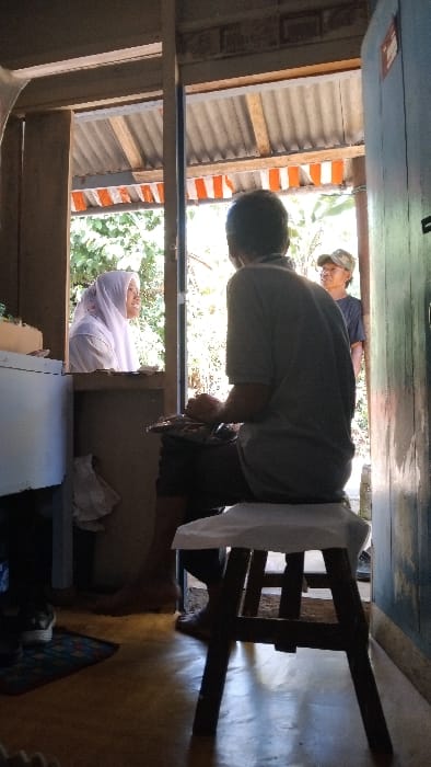 A man sits on a wooden stool with a white cushion, facing a woman wearing a white headscarf. The woman is standing in a doorway. The man is wearing a grey t-shirt and black shorts. His feet are bare. The woman is wearing a long white dress and has her hair covered with the headscarf. The man is looking at the woman, and she is looking back at him. The man's right leg is extended, and his left leg is bent and resting on the stool. The woman has a white bag on her shoulder. Behind the man, in the doorway, is another man wearing a grey t-shirt and a blue and white hat.  The man is standing and facing the woman. The scene takes place in a doorway between a room and a porch. The doorway has a corrugated metal roof with orange and white stripes. The room is sparsely furnished with a white refrigerator in the corner, and a small table. The floor of the room is a light brown wood, while the porch is made of concrete and is covered with vegetation. The photograph appears to have been taken in Kecamatan Panjalu, Indonesia, at the coordinates -7.143, 108.278.  The light is coming from the left side of the room, creating a slight shadow on the right side of the scene. The room is well-lit and the man is in focus. The woman and the other man are blurred in the background.  The photograph is slightly tilted.