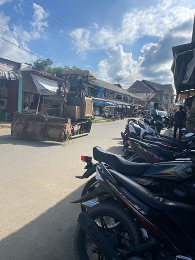 In Muliengmanyang, Indonesia, a bulldozer is seen on an asphalt road in front of a row of shops. The bulldozer, partially covered in dirt and mud, has a large bucket in the front. Several motorcycles are parked on the side of the road, some in front of shops and others on the sidewalk. Two men are visible in the background, one standing and one walking. The sky is partly cloudy with a bright sun, casting shadows on the road. The buildings in the background appear to be a mix of traditional and modern structures.