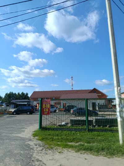 The image, captured on a sunny day, shows a street scene in the city of Snopok Stary, Russia. A light blue sky with scattered white clouds dominates the upper portion of the frame, with power lines crisscrossing overhead. In the middle ground, a long, single-story building with a reddish-brown tiled roof is visible, featuring several large windows and a brick facade. Several cars are parked in front of the building, including a dark blue minivan and a dark sedan. A green metal fence runs along the edge of the property, with a sign displaying operating hours: "8-18", "8-19", and "8-17". Beyond the building, a cellular tower stands tall against the sky. The foreground consists of a paved road with some dirt and patchy vegetation. The scene appears to be a commercial area, possibly a market or a business complex. There are no people clearly visible in the image.