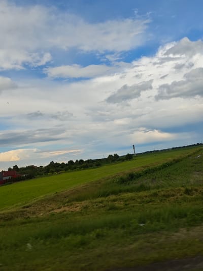 The image depicts a rural landscape under a cloudy sky, likely within the Красненскі сельскі Савет, Belarus. In the foreground, a grassy field slopes upwards and to the right, with visible textures of dried grass and vegetation. Further up the slope, a larger expanse of green field stretches across the middle ground. On the left side of this green field, a red-roofed building with a white facade and a row of red fencing is visible amongst trees. In the background, a line of trees forms a distant horizon, punctuated by a tall, slender structure resembling a water tower or radio mast. The sky is a mix of blue patches and white and gray clouds, with a bird in flight visible in the upper left portion of the image. The overall impression is one of a peaceful, agricultural setting.