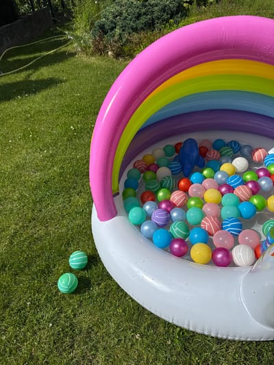 An inflatable rainbow-themed ball pit is situated on a well-maintained green lawn. The ball pit contains numerous colorful plastic balls of various sizes and hues, including pink, blue, yellow, green, purple, and white, some with striped patterns. A small blue inflatable dolphin toy rests among the balls. Two green and white striped balls are lying on the grass outside the ball pit. The scene is illuminated by bright sunlight, casting shadows on the lawn. The background shows greenery, possibly bushes and trees, and a portion of a hosepipe. The location is identified as the city of Astrashitskagaradokski rural council, Belarus. There are no people visible in the image.