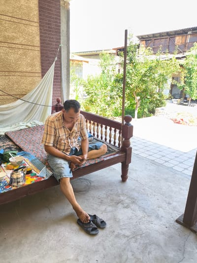 In Paytugh, Uzbekistan, a man sits on a traditional low wooden daybed, his legs extended, one resting on the concrete floor. He wears a plaid short-sleeved shirt and denim shorts. His feet are bare, with dark sandals placed in front of him. On the daybed, next to him, is a collection of items including a stainless steel teapot, bowls, and what appears to be a tea set, arranged on patterned cloths. The daybed has a dark wooden frame with decorative spindles. Behind the daybed, a light-colored draped fabric hangs against a textured wall. The setting is an outdoor, covered area, possibly a veranda or patio, with a tiled floor extending to the right. Lush green trees and foliage are visible in the background, along with parts of a building. The scene is lit by natural daylight, casting shadows on the concrete floor.