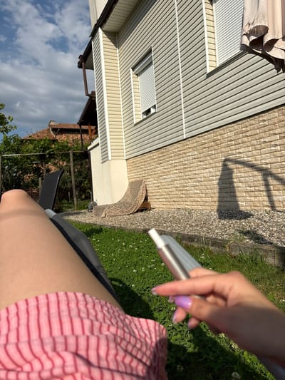 A first-person perspective image depicts an individual reclining outdoors in Polena, Bulgaria, under a partly cloudy sky. The subject's bare legs and a portion of a pink and white striped garment are visible in the foreground, resting on a dark grey outdoor lounger. The subject's right hand, featuring purple-tipped nail polish, holds a sleek, metallic silver electronic device, possibly a vape pen. The immediate ground consists of green grass with white clover flowers, transitioning to a concrete curb and then an area of light-colored gravel.
In the mid-ground, a two-story residential building features light beige horizontal siding on its upper facade and a light-colored brick veneer foundation. Multiple windows with white roll-down shutters are present, with one shutter partially open. Brown gutters and a downspout are affixed to the building's exterior. Strong sunlight creates distinct shadows, including a human-like silhouette, cast upon the brick foundation and gravel. To the right of the building, a partially furled beige umbrella is visible. Further in the background, dense green foliage, including trees, and the reddish-brown tiled roof of another structure are discernible. A patterned fabric object is casually leaned against the house near the foundation level.