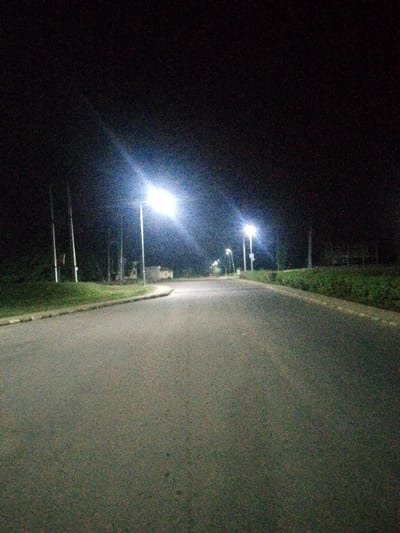 A nighttime view shows an asphalt road curving into the distance, illuminated by intermittent streetlights. Dark hedges or vegetation flank both sides of the road. The sky is a featureless black. Several light poles are visible along the roadside, contrasting with the dark sky. The road surface appears smooth, and the overall scene evokes a sense of quiet isolation.