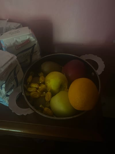 The image shows a still life arrangement on a dark wooden surface, likely a table, against a pale pink wall. In the foreground, a metal pot contains several fruits and candies. The fruits include what appear to be two apples (one green, one yellowish-green), a red apple, and an orange. Scattered among the fruit are several individually wrapped yellow candies.
Behind the pot, to the left, are two large bags of what appears to be flour. The bags have labels on them, one clearly showing "CORTICELLA" in red lettering, along with text that includes "Farina" and what looks like a product code and expiration date "10.12.2026". The other bag is partially obscured but also appears to be a food product with text on it.
The lighting in the image is dim, with a light source casting shadows and highlighting certain areas, particularly the fruits in the pot and the packaging of the flour bags. The overall atmosphere is that of an indoor setting, possibly a kitchen or pantry. There are no people or animals present in the image. The scene is static and composed.
The location of Mussolini, Italy is not discernible from the image.